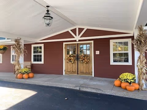 Entrance of a building with double wooden doors decorated with autumn wreaths, flanked by windows. The exterior walls are painted red with white trim. There are pumpkins and potted yellow flowers on either side of the entrance, along with cornstalk decorations. A covered driveway with a hanging lantern light fixture is visible.