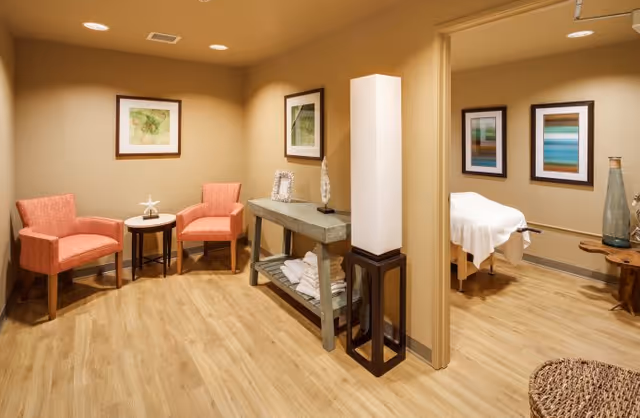 Warm-toned interior relaxation area with two coral chairs, a console table and a doorway to a treatment room with a covered massage table.