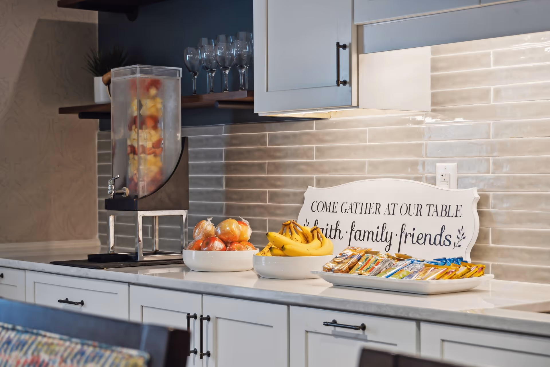 A kitchen counter with a beverage dispenser filled with fruit-infused water, bowls of apples and bananas, and a tray of assorted snack bars. Behind the counter is a decorative sign that reads 'COME GATHER AT OUR TABLE faith family friends' and a backsplash of light gray subway tiles. There are white cabinets above and below the counter and a shelf with wine glasses.