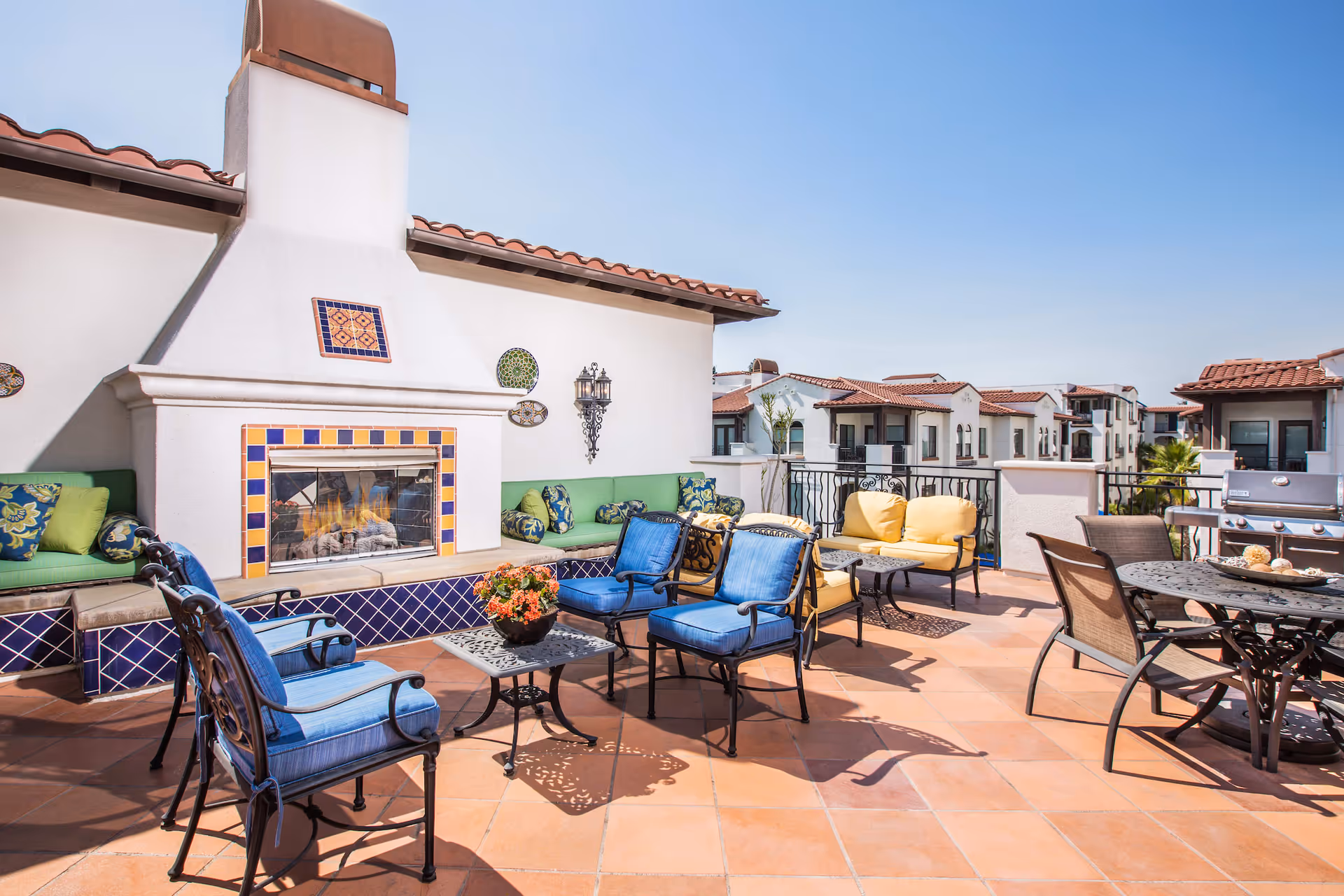 Outdoor patio area with a tiled fireplace, cushioned seating in blue, green, and yellow, a small table with a flower pot, a round dining table with chairs, and a barbecue grill under a clear blue sky with surrounding buildings in the background.