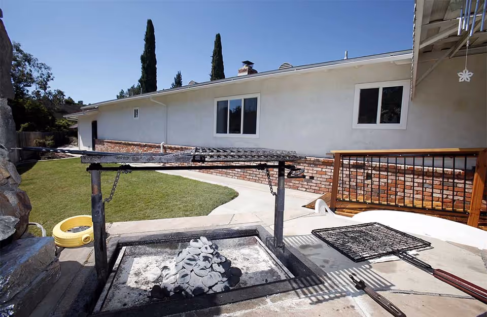 Outdoor barbecue grill area with charcoal briquettes on the grill, a metal cooking grate, and a white building with windows and a brick lower wall in the background under a clear blue sky.