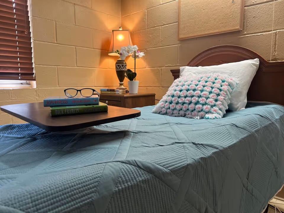 A neatly made single bed with a teal quilted bedspread and a decorative pillow with pink and white flowers. On the bed is a wooden tray holding two books and a pair of black-rimmed glasses. Next to the bed is a wooden nightstand with a table lamp, a small potted orchid, and additional books. The room has beige brick walls and a window with closed wooden blinds.