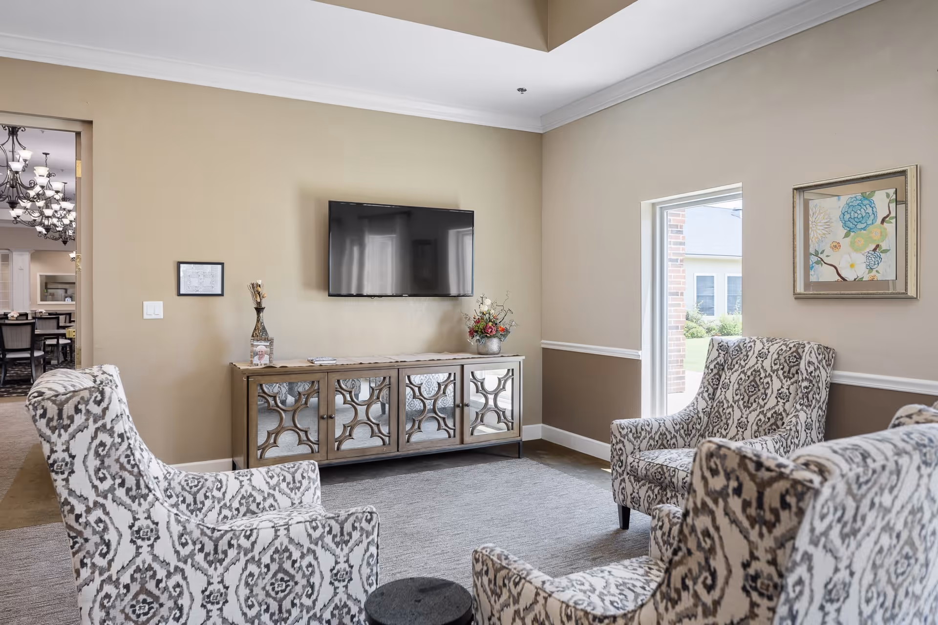 A cozy living room with four patterned armchairs arranged around a small black round table. A flat-screen TV is mounted on the beige wall above a decorative wooden cabinet with mirrored doors. There is a small window letting in natural light and a framed floral artwork on the wall. Through an open doorway, a dining area with chandeliers and tables is visible.