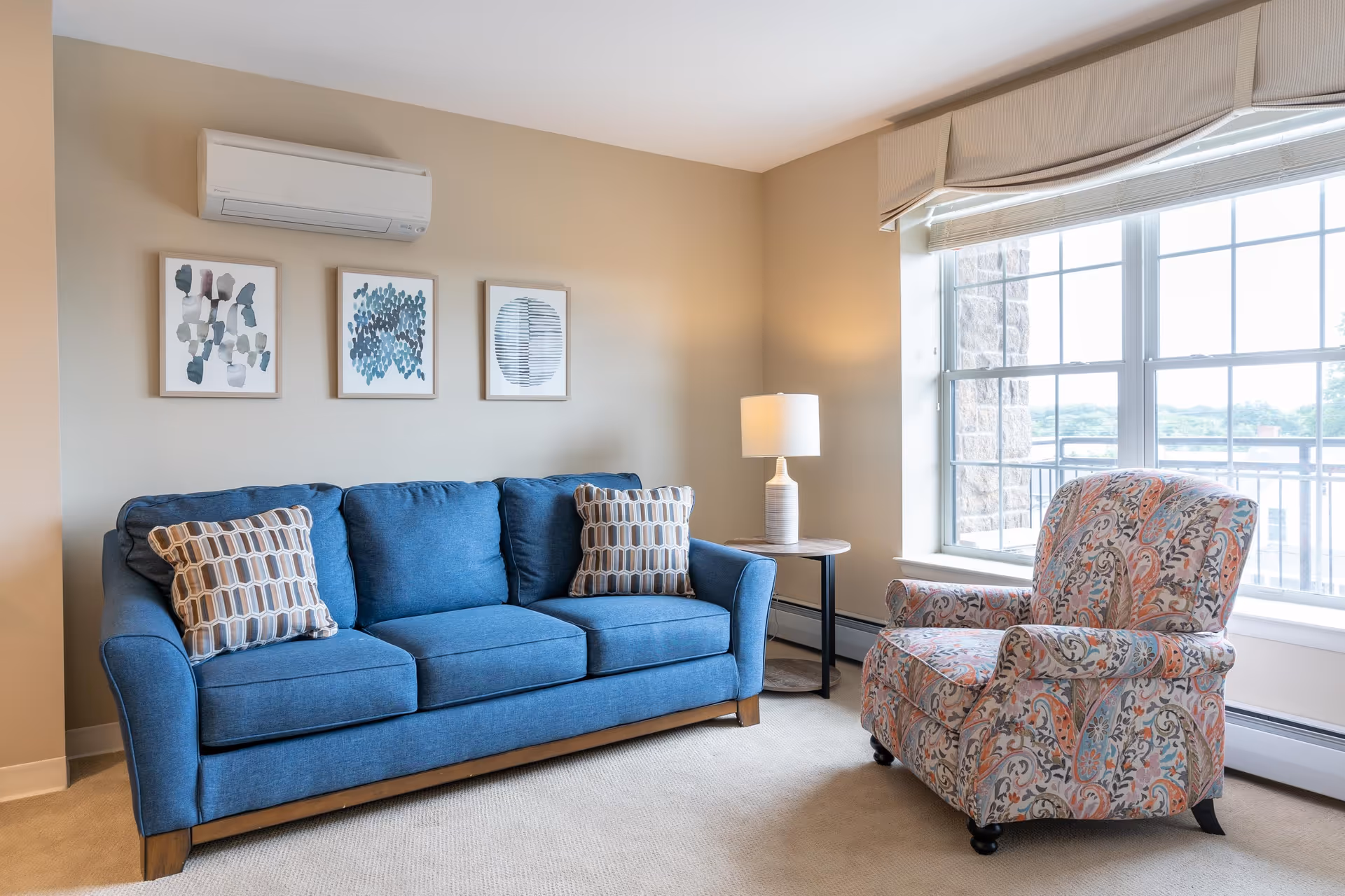 A cozy living room with a blue three-seater sofa adorned with two patterned pillows, a colorful patterned armchair, a round side table with a white lamp, three abstract art pieces on the beige wall, and a large window with a view outside.