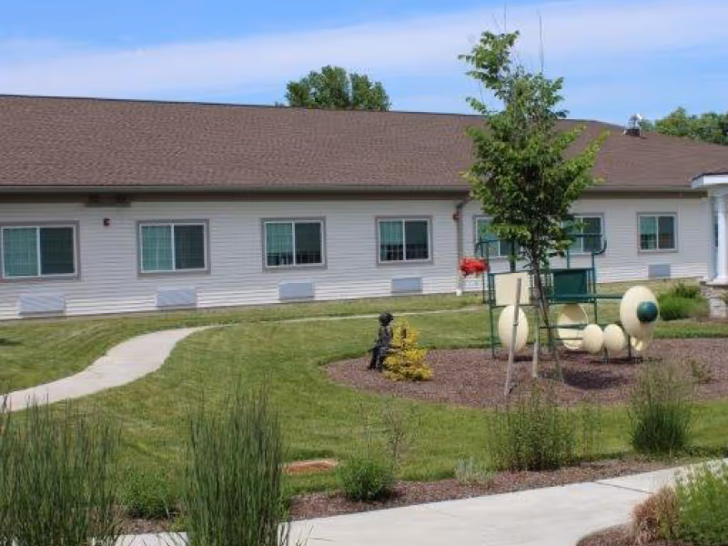 Outdoor view of a single-story building with a brown roof and white siding. In front of the building is a well-maintained lawn with a curved concrete pathway, a small tree, and a whimsical caterpillar-shaped bench sculpture. There are some bushes and plants along the walkway.