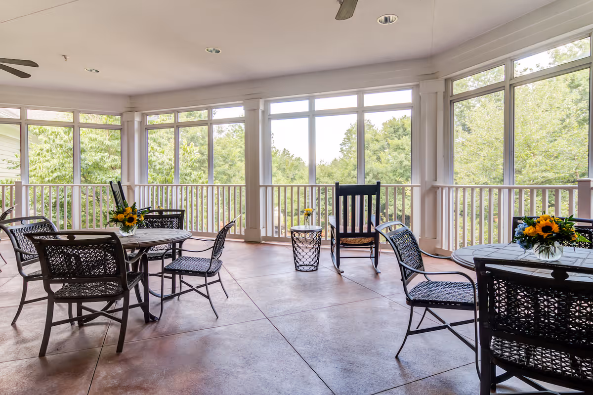 A spacious screened-in porch area with multiple metal tables and chairs arranged for seating. Each table has a small vase with sunflowers. There is also a black rocking chair and a small round side table with a flower vase. Large windows surround the porch, offering a view of green trees outside.