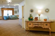 A well-lit hallway in a senior living facility with a decorative wooden console table on the right, adorned with a lamp, floral arrangement, and decorative items. In the background, there is a seating area with two blue armchairs and a window with curtains.