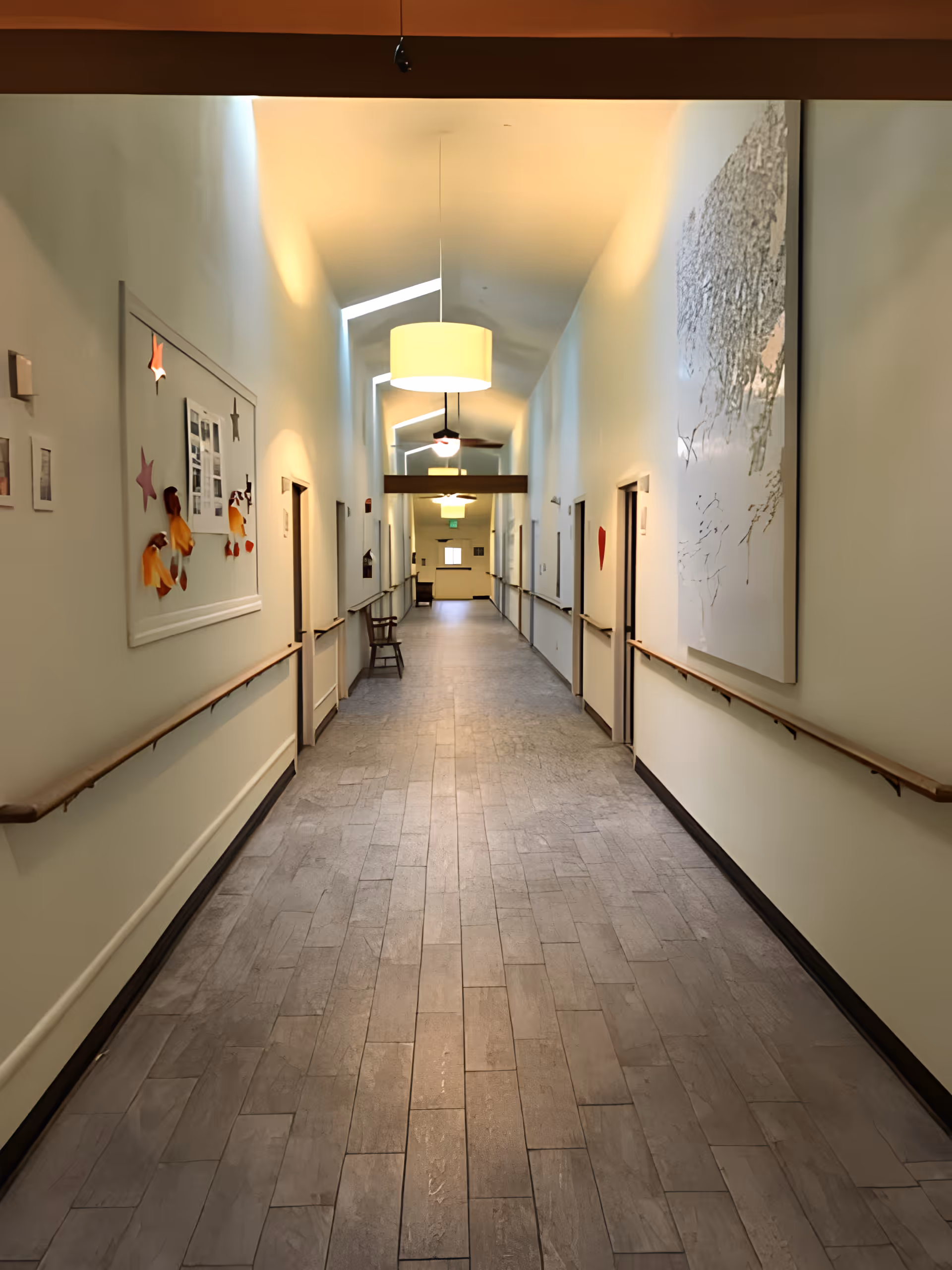 Long, well-lit interior hallway in an assisted living facility with handrails, doors on both sides, artwork on the walls and overhead lighting.