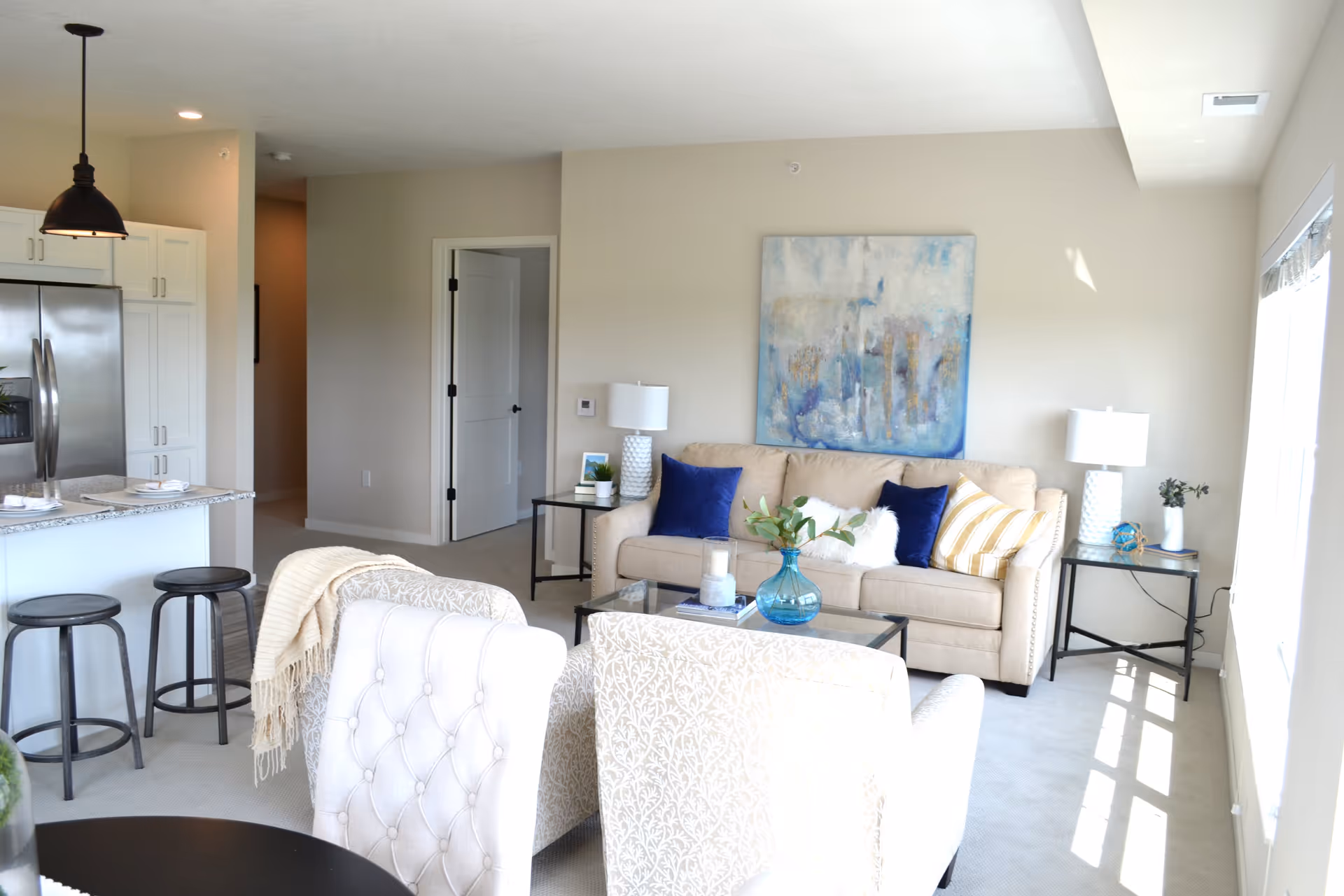 Bright and modern living room area with a beige sofa adorned with blue and striped pillows, two patterned armchairs, a glass coffee table with a blue vase and candles, side tables with lamps, and a large abstract painting on the wall. The room is adjacent to a kitchen area with a granite countertop, three black bar stools, white cabinets, and a stainless steel refrigerator. Sunlight streams in through a large window on the right.