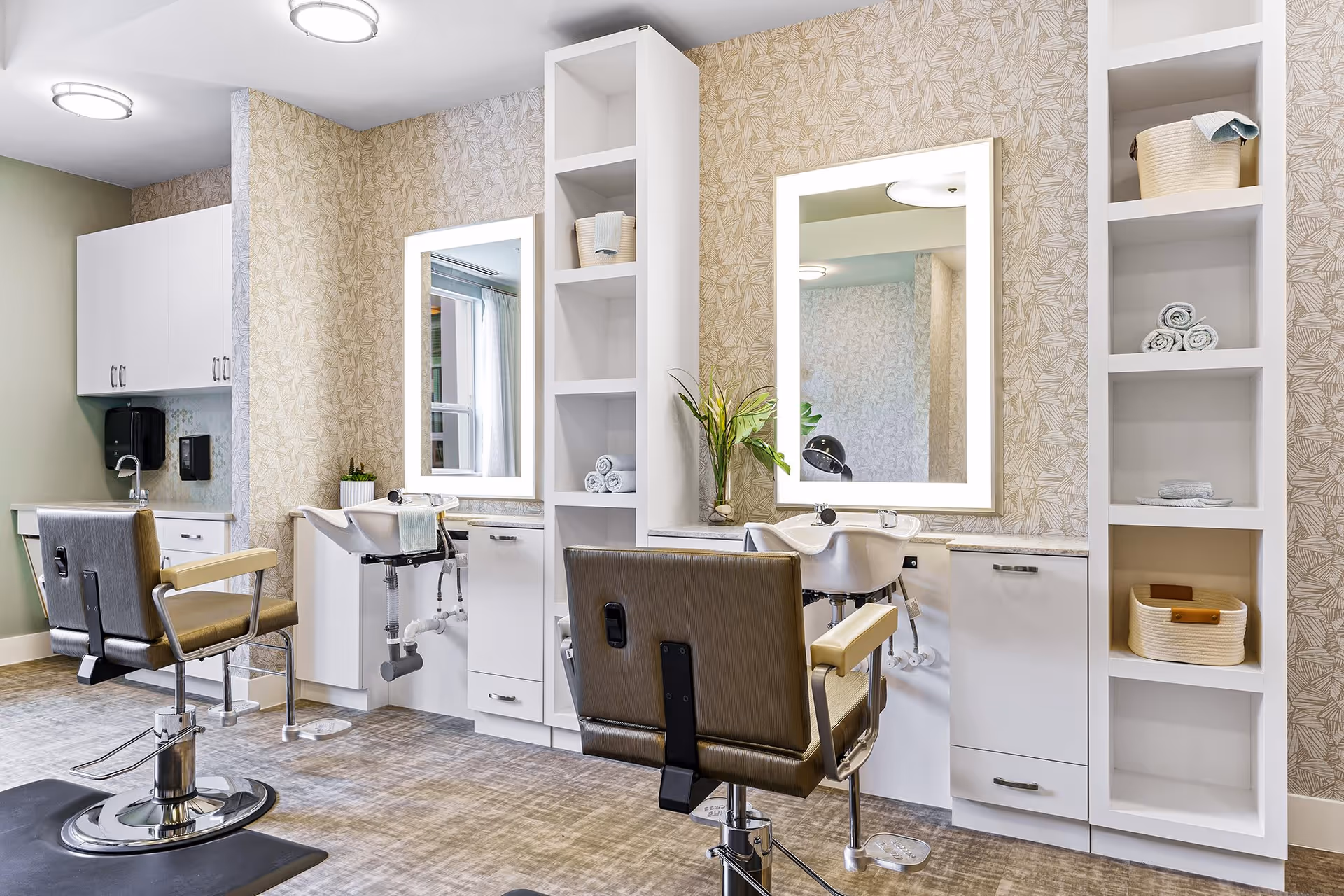 Bright salon-style grooming area with two styling chairs, backwash sinks, illuminated mirrors, and open shelving.