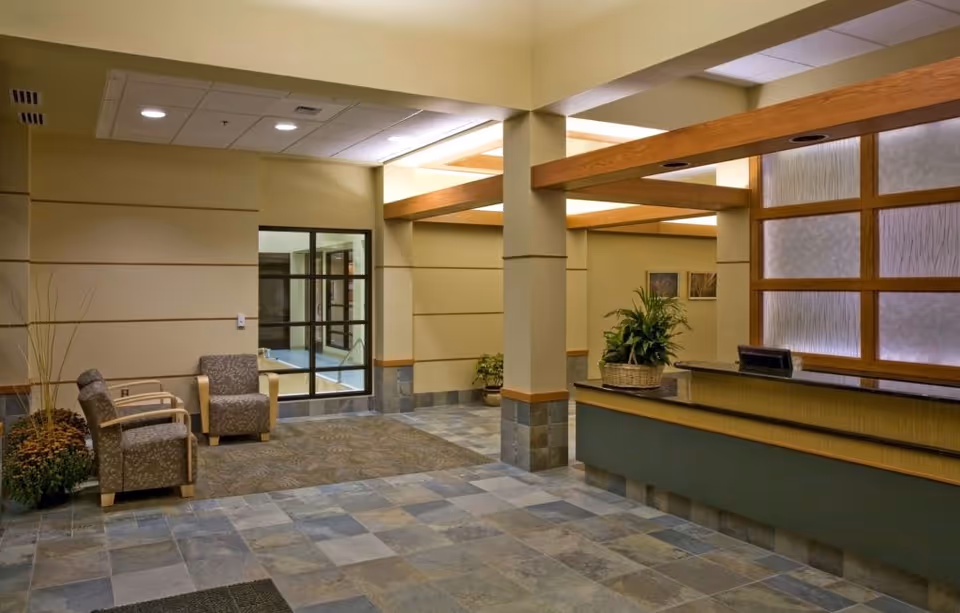 Reception area of a senior living facility with a front desk on the right, decorated with a basket of plants. To the left, there are two patterned armchairs next to a window and some potted plants. The floor is tiled with a mix of gray and beige tiles, and the walls are painted in a light beige color with wooden accents on the ceiling and around the reception desk.