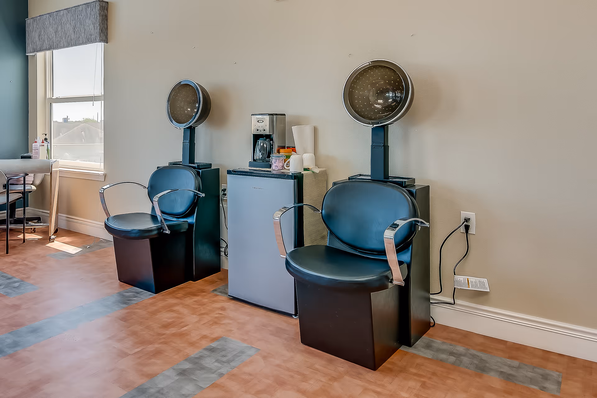 Interior view of a salon area in a senior living facility featuring two black hair drying chairs with overhead hair dryers, a small refrigerator, a coffee maker, and a window with a gray valance letting in natural light.