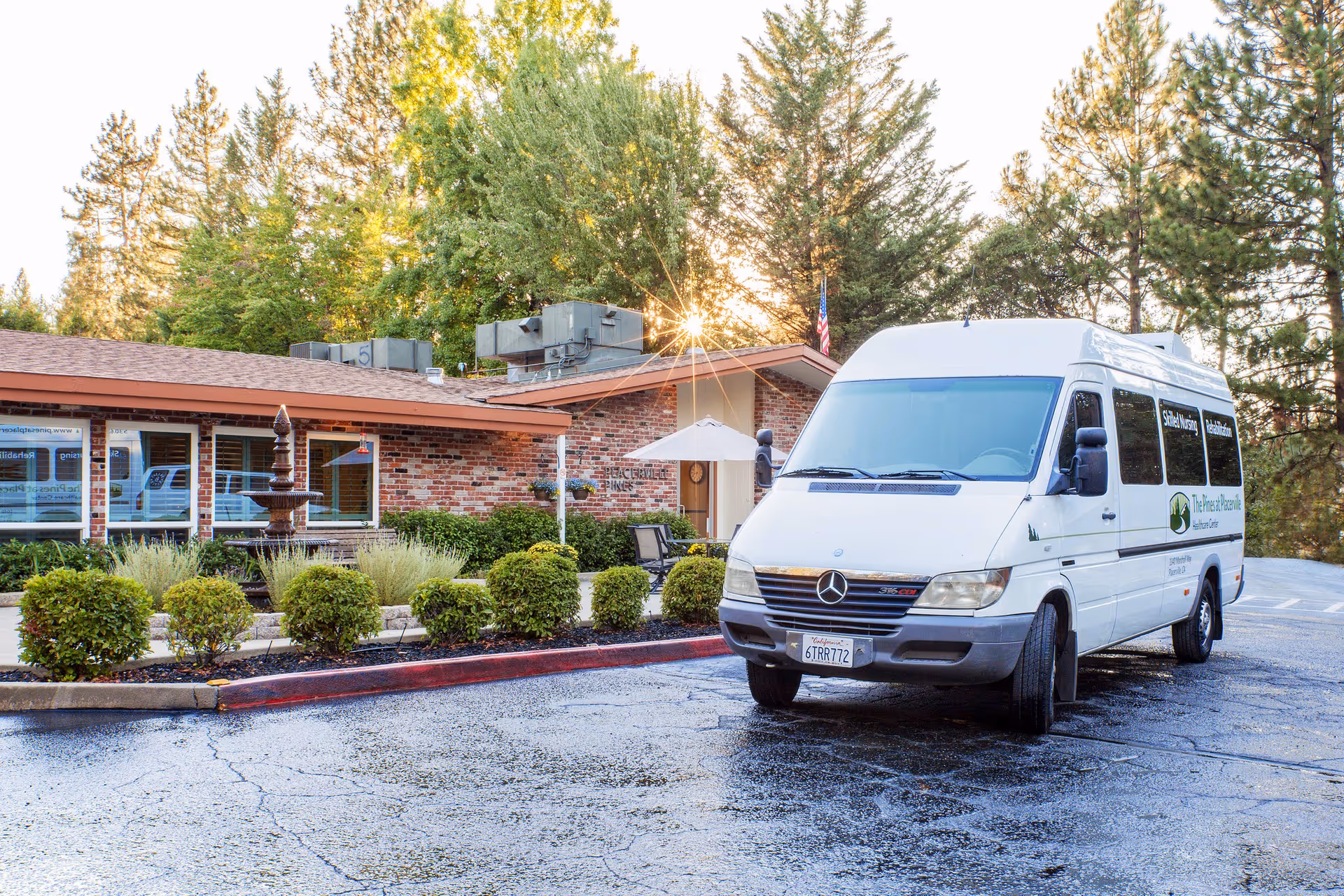 A white Mercedes van parked in front of a brick building with a red roof, surrounded by green bushes and trees. The building has a small fountain and outdoor seating with an umbrella. The sun is shining through the trees in the background.