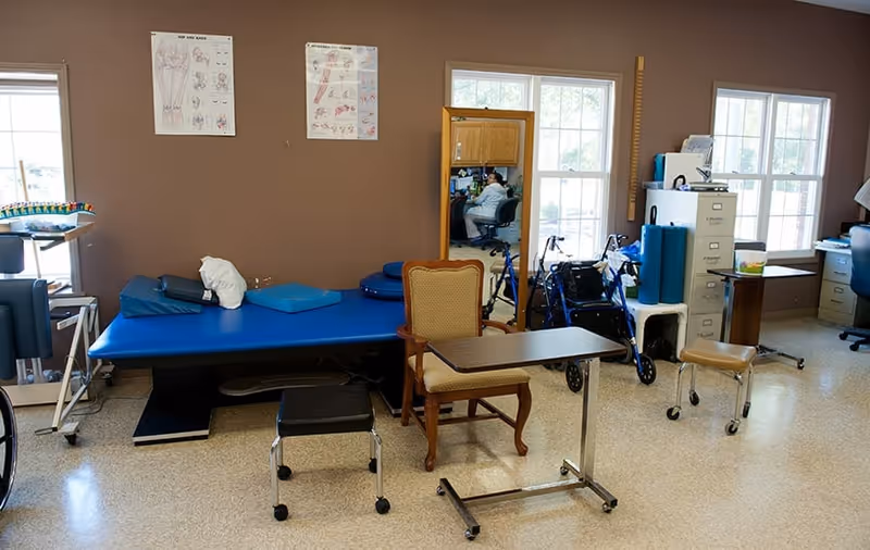 A rehabilitation therapy room with a blue treatment table, chairs, walkers and medical equipment against a brown wall with windows.