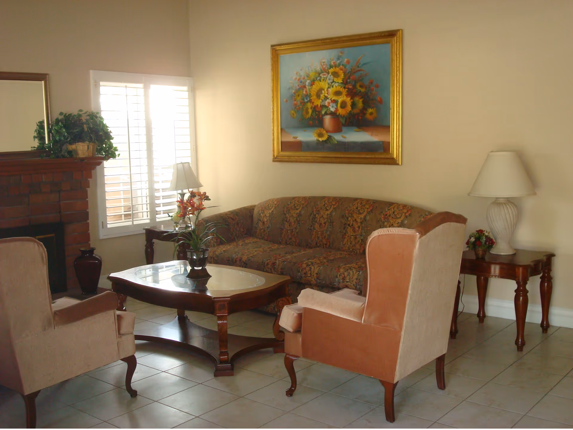A cozy living room with a patterned sofa, two upholstered armchairs, a wooden coffee table with a glass top, a side table with a lamp, a brick fireplace with a plant on the mantel, and a large framed painting of sunflowers on the wall.