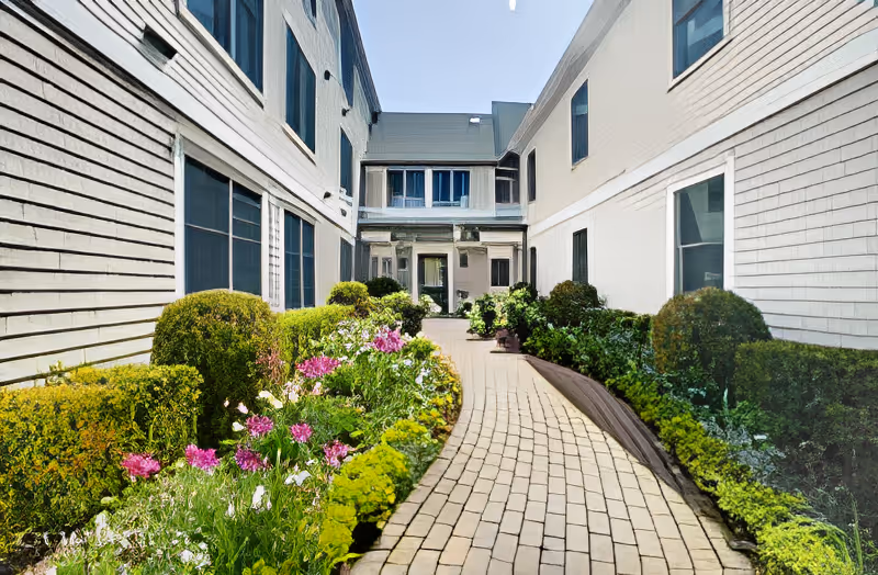 Brick walkway through a landscaped courtyard between two white-sided buildings leading to an entrance.