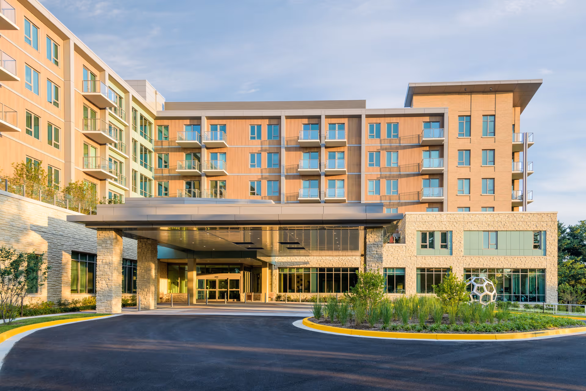 Exterior view of The Seneca Rockville senior living facility showing a modern multi-story building with balconies, large windows, a covered entrance, and landscaped greenery in front under a clear sky.