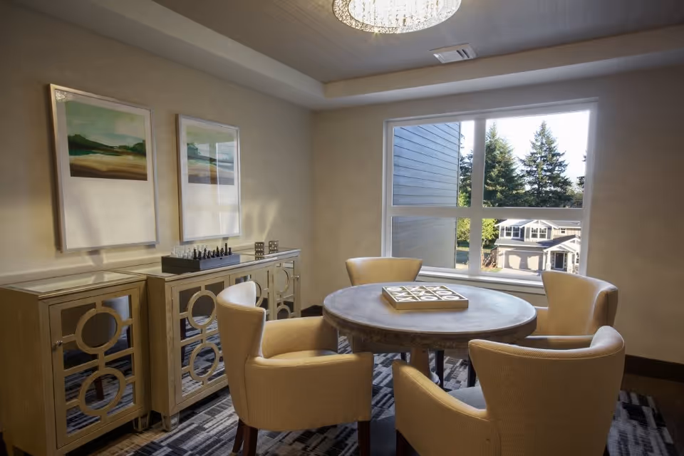 A cozy interior room with a round wooden table surrounded by four beige upholstered chairs. On the table is a tic-tac-toe game board. Behind the table is a mirrored cabinet with a chess set and two framed landscape paintings on the wall above it. A large window shows a view of trees and houses outside. A chandelier hangs from the ceiling.