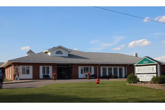 Exterior view of Ambassador Health of Nebraska City, a single-story brick building with a gray shingled roof, white-framed windows, and a green sign on the right side displaying the facility's name and address. The building is surrounded by a well-maintained lawn under a blue sky with some clouds.