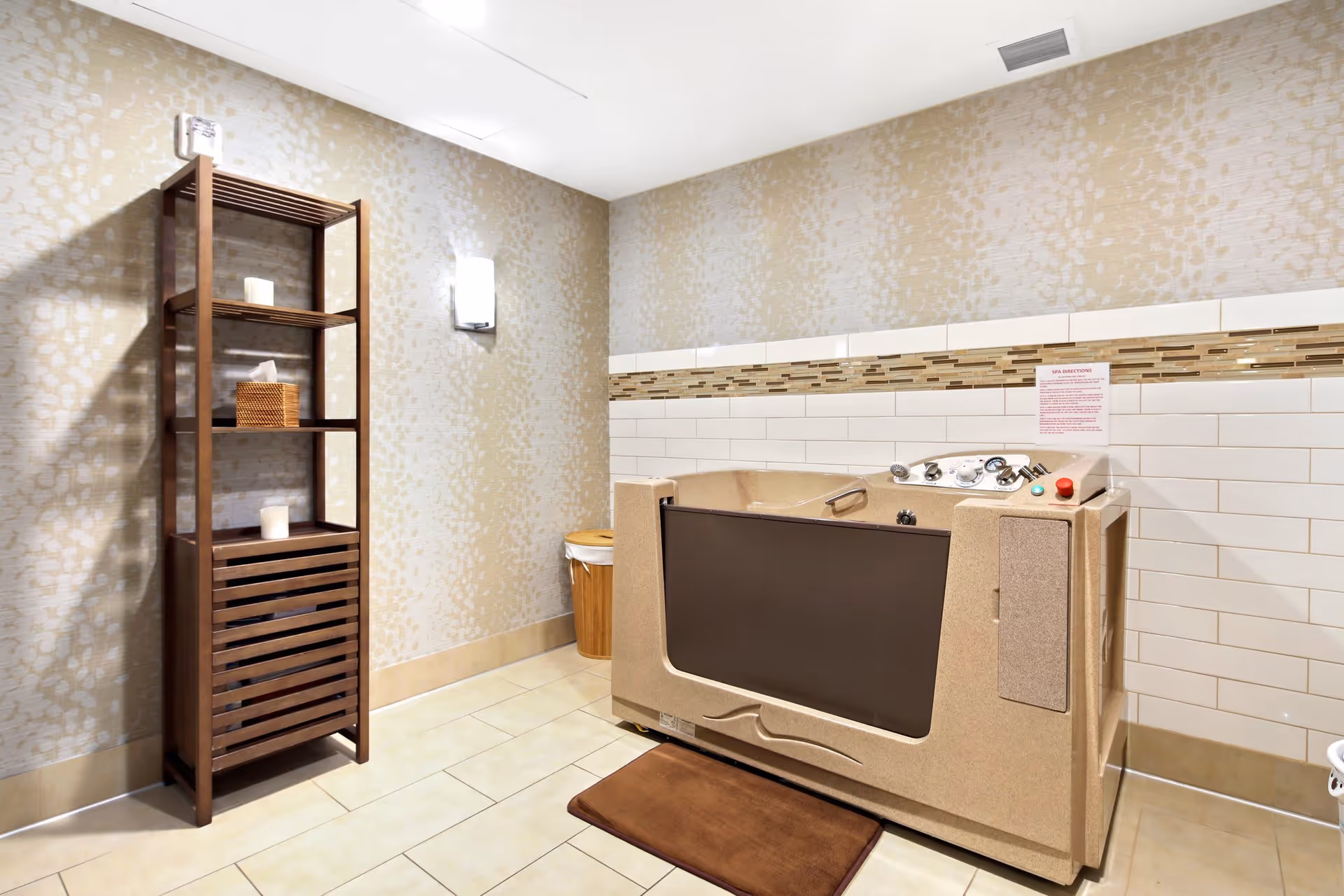A beige walk-in bathtub with control knobs and a safety handle is positioned against a tiled wall with a decorative mosaic strip. To the left, there is a tall wooden shelving unit holding candles and a tissue box. The floor is tiled, and a brown bath mat is placed in front of the tub. The walls have a light patterned wallpaper, and a wall-mounted light fixture is visible above the shelving unit.