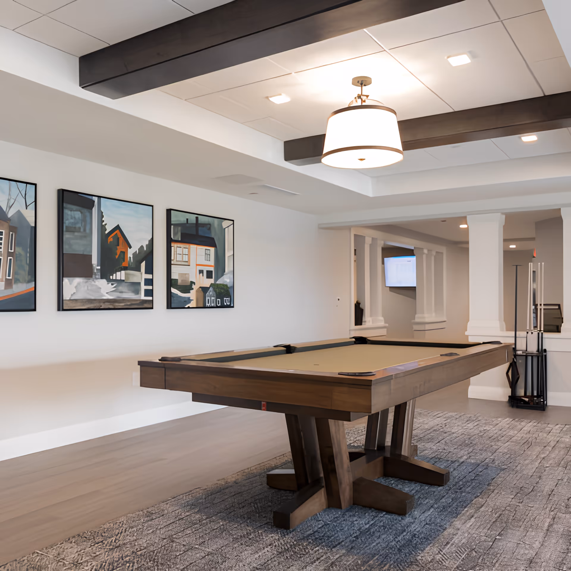 A modern game room featuring a wooden pool table with a beige felt surface, positioned on a textured gray rug. The room has white walls adorned with three framed paintings depicting houses and trees. The ceiling has dark wooden beams and a large round light fixture. In the background, there are white columns and a rack holding pool cues.