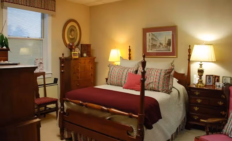 A cozy bedroom with a wooden four-poster bed dressed in white bedding, plaid pillows, and a burgundy throw blanket. The room features two wooden nightstands with lamps, a wooden dresser, a chair with a red cushion, framed pictures on the walls, and a window with blinds and a valance.