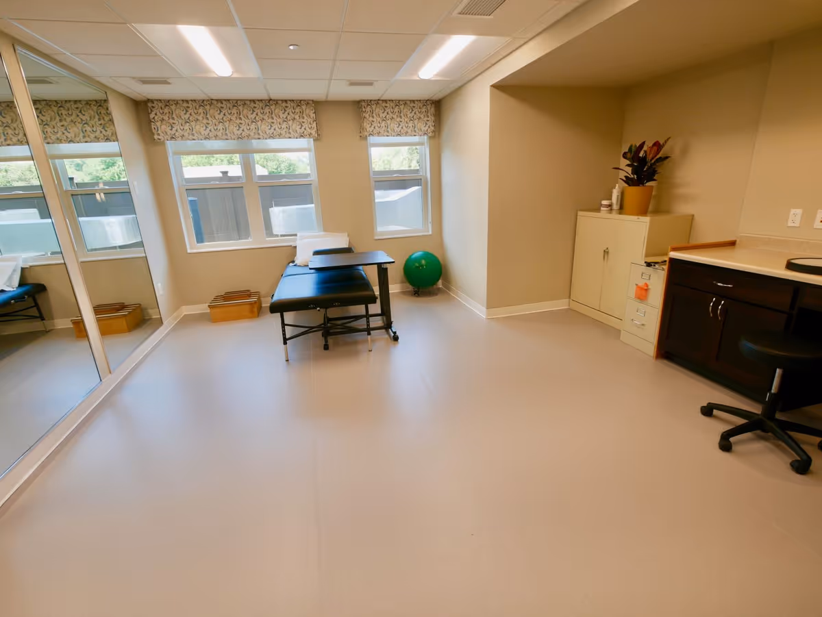 A bright therapy/exercise room with a treatment table, green exercise ball, mirrored wall, cabinets and a desk.