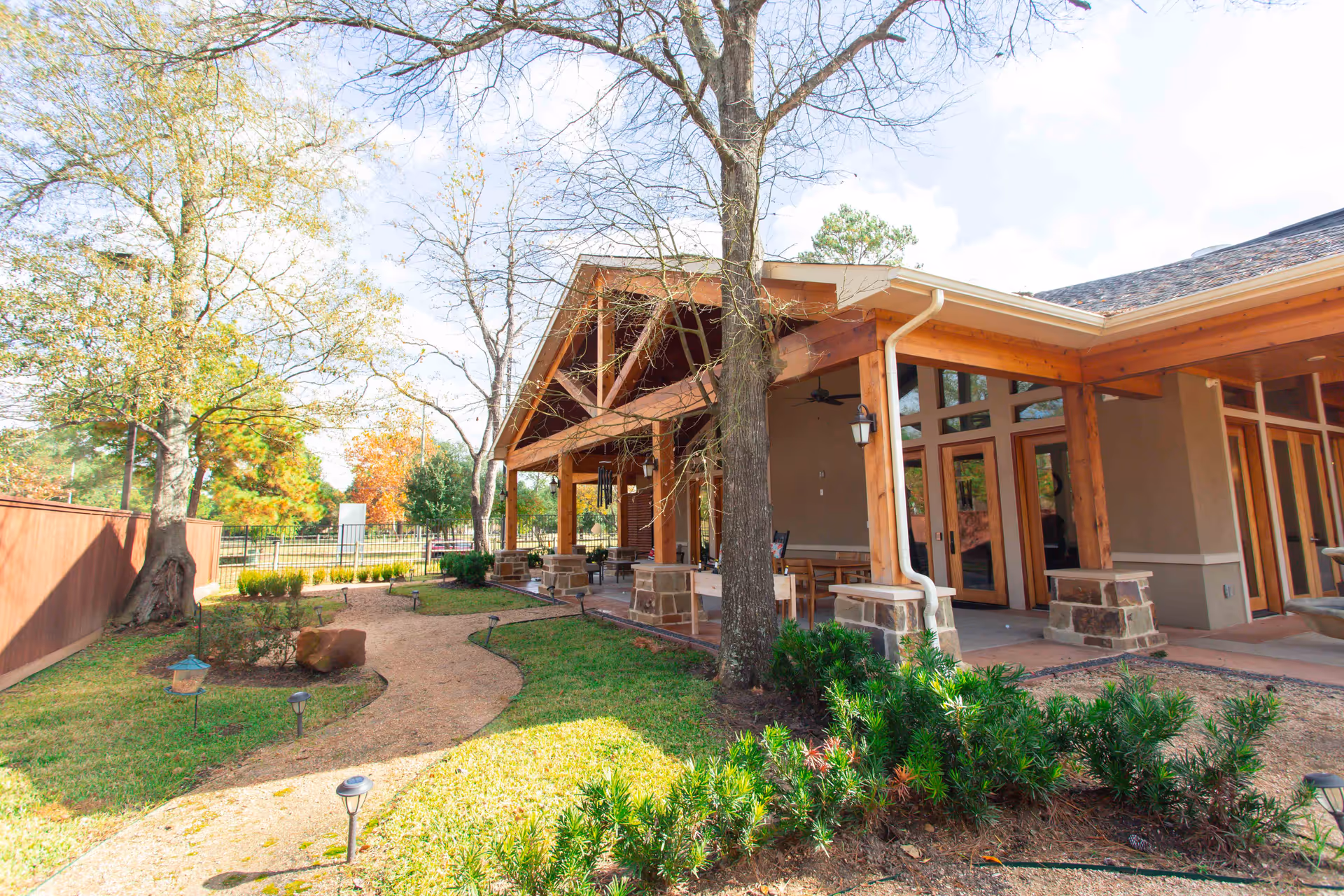 Covered outdoor patio with wooden beams and seating beside a landscaped walkway and garden next to a single-story building.