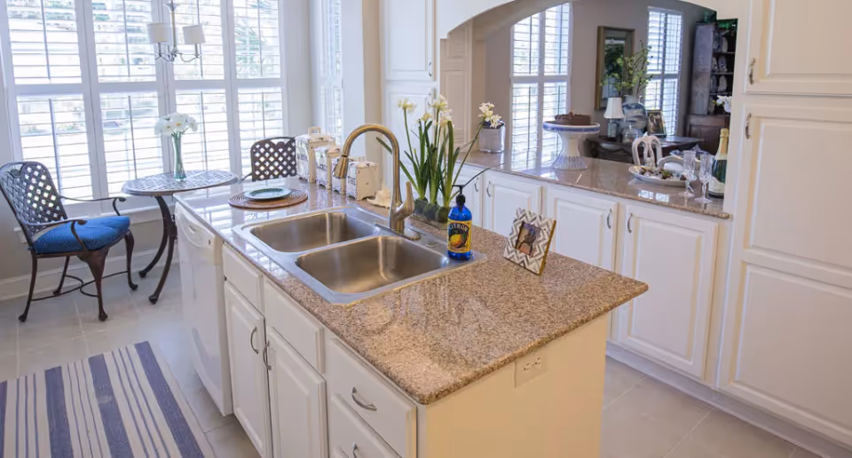 Bright kitchen with white cabinets and granite countertops featuring a double sink with a gold faucet. A small round table with two chairs is placed near large windows with white shutters, allowing natural light to fill the space. Decorative items including flowers, a framed photo, and kitchen accessories are arranged on the counters.
