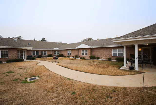 Outdoor courtyard area of a senior living facility with a paved walkway, dry grass, bushes along the building walls, and a covered patio with chairs and tables.