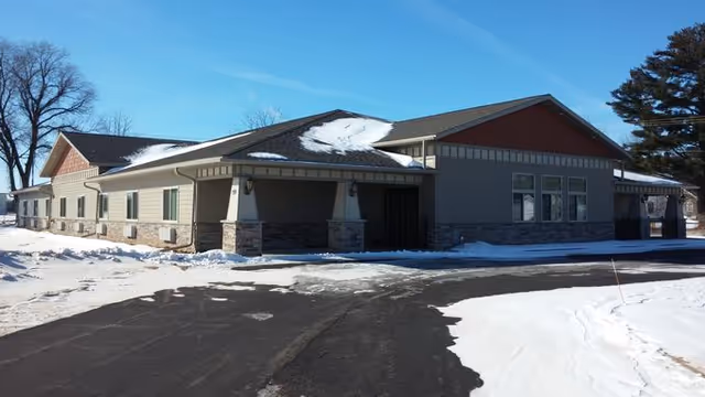 Exterior view of a single-story building with a sloped roof partially covered in snow. The building has a combination of stone and siding on the walls, with several windows and a covered entrance. Snow covers the ground around the building and the sky is clear and blue.
