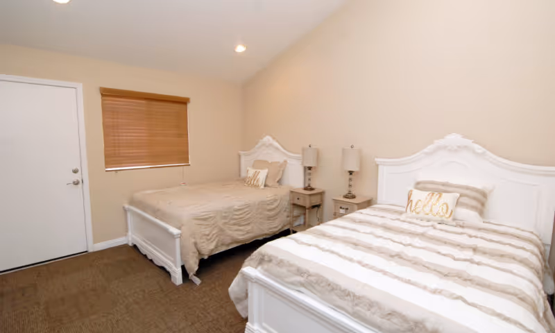 A bedroom with two white wooden beds, each with beige and white bedding and a decorative pillow that says 'hello'. There are two matching bedside tables with lamps between the beds. The room has beige walls, a brown carpet, a window with closed wooden blinds, and a white door.
