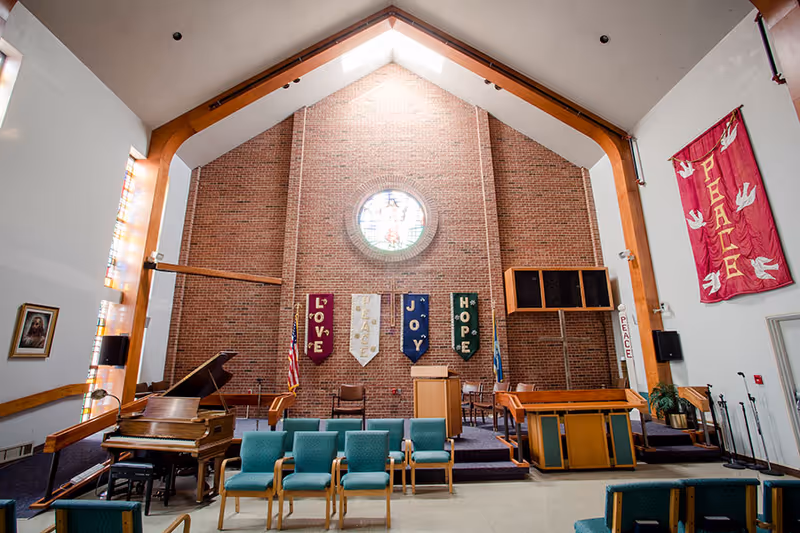 Interior view of a chapel or worship area with a high vaulted ceiling and a large circular stained glass window on a brick wall. The room features a piano on the left, several rows of green cushioned chairs, a wooden podium, and banners hanging on the wall with the words LOVE, PEACE, JOY, and HOPE. There is also a large red banner with the word PEACE on the right wall and an American flag near the podium.