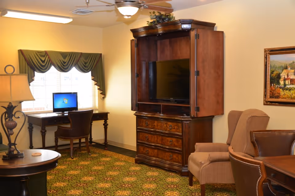 A cozy interior room with a wooden desk and chair in front of a window with green drapes, a computer on the desk, a wooden cabinet with an open door revealing a flat-screen TV, a beige armchair, a round table with a lamp, and a framed painting on the wall.