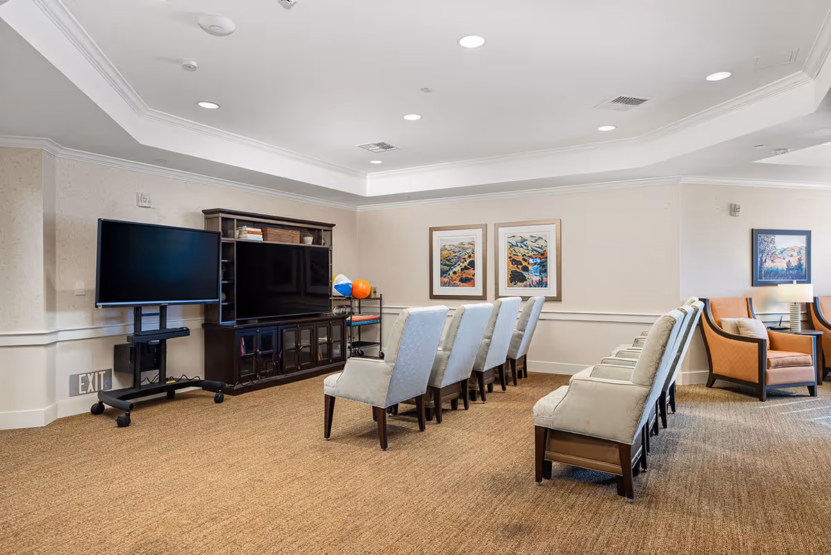 A spacious common room with two rows of beige upholstered chairs facing a large flat-screen TV on a dark wooden cabinet. Another TV on a mobile stand is positioned to the left. The room has beige walls with white trim, carpeted floor, recessed ceiling lights, and framed landscape artwork on the walls. Two orange armchairs with cushions and a side table with a lamp are visible in the background.