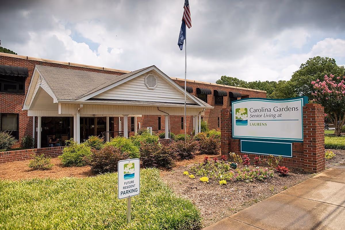 Exterior view of Carolina Gardens at Laurens senior living facility showing the entrance with a covered porch, landscaped garden with flowers and shrubs, an American flag on a flagpole, and a sign indicating the facility name and future resident parking.