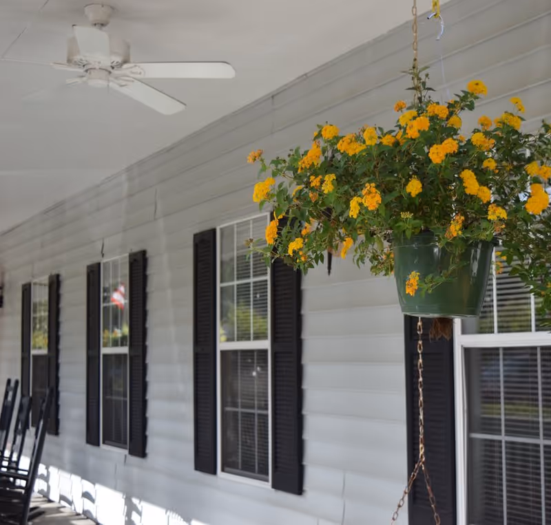 Covered porch with a hanging pot of yellow flowers, white siding, black shutters, windows and rocking chairs.
