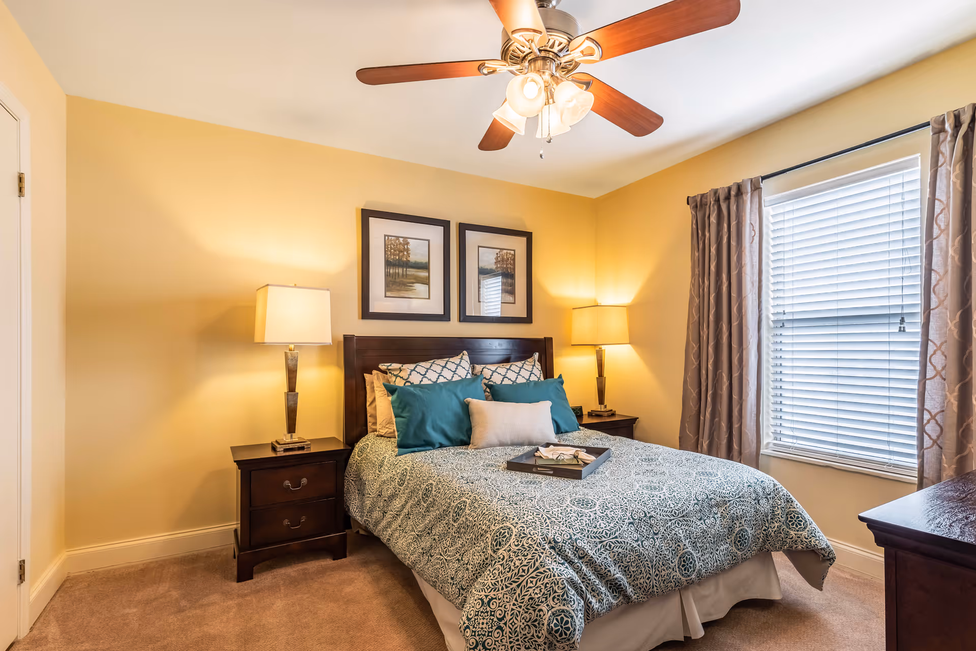 A cozy bedroom with a double bed featuring a patterned blue and white bedspread and multiple pillows. There are two dark wooden nightstands on either side of the bed, each with a lamp. Above the bed, two framed landscape paintings hang on the yellow walls. A window with blinds and patterned curtains is on the right side, and a ceiling fan with lights is mounted on the ceiling.