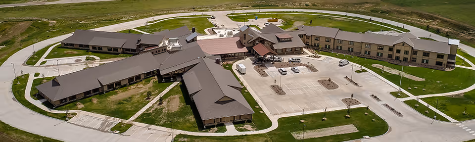 Aerial view of The Ranch House senior living facility showing multiple connected buildings with brown roofs, surrounded by paved roads and parking lots, set in a green grassy area.