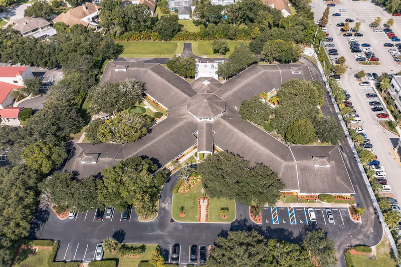 Aerial view of The Landmark senior living facility showing a large building with a unique X-shaped roof surrounded by trees, parking lots, and nearby residential houses.