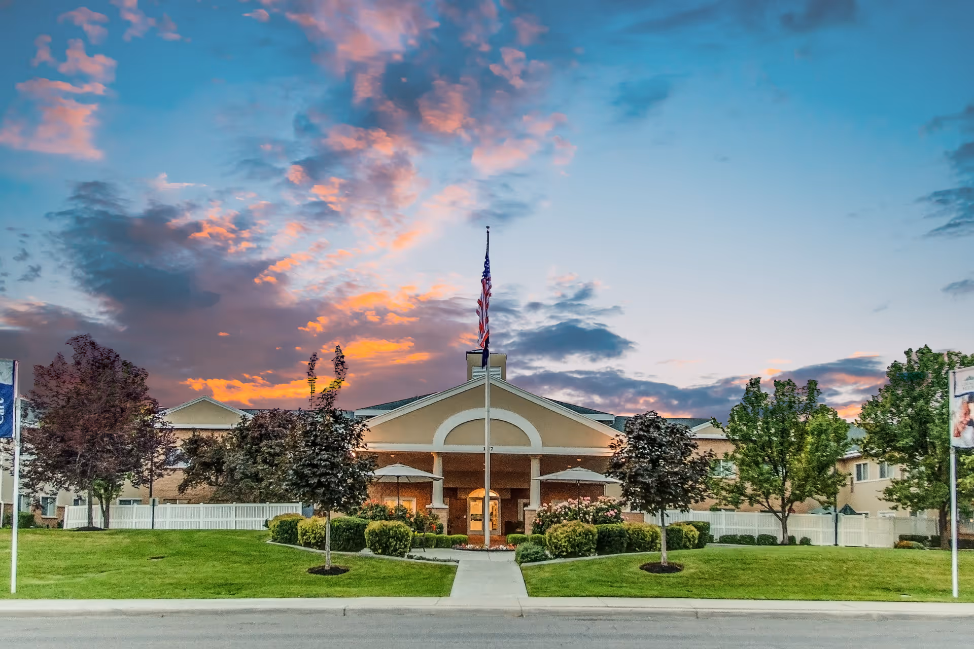 Front exterior view of Legacy House of South Jordan building at sunset with a flagpole in the center, surrounded by green lawns, trees, and shrubs.