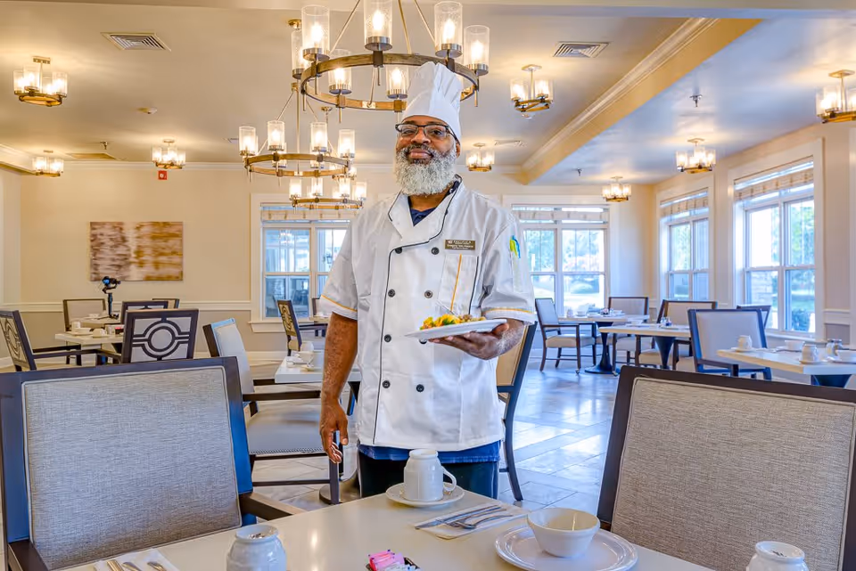 A chef wearing a white uniform and hat stands in a bright dining room holding a plate of food. The room has multiple tables and chairs, large windows letting in natural light, and modern chandeliers hanging from the ceiling.