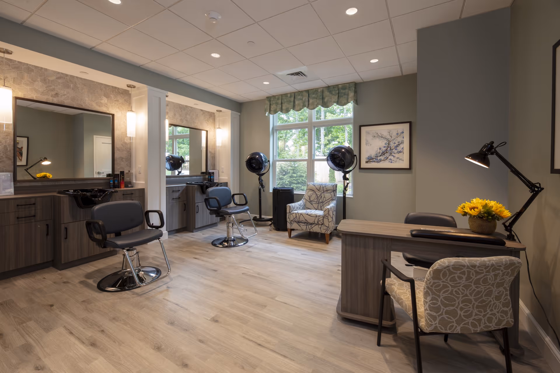 Interior of a salon area in a senior living facility with two salon chairs in front of mirrors and sinks, two hair dryers on stands near a window with green valance, a patterned armchair, and a desk with a lamp and a vase of yellow flowers.
