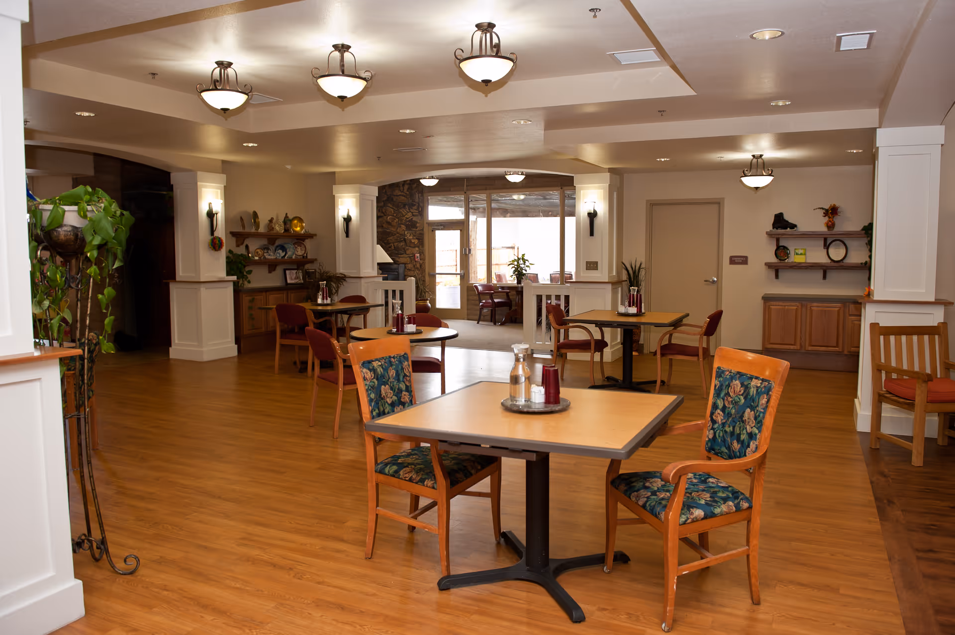 A spacious dining area in a senior living facility with wooden floors, several tables and chairs with floral upholstery, and decorative plants. The room is well-lit with ceiling lights and wall sconces, and there is a doorway leading to another room with more seating visible.