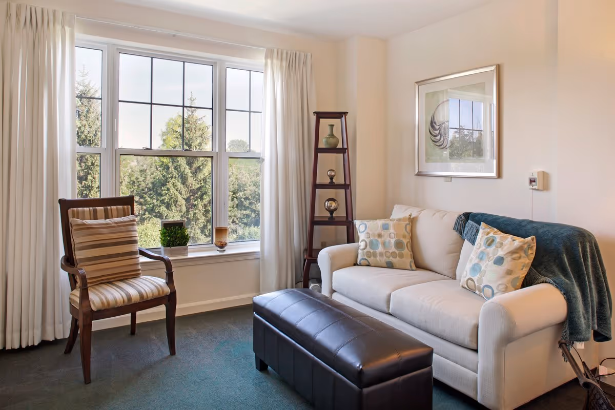 Cozy living room with a sofa, armchair, leather ottoman, tall window and decorative shelving.