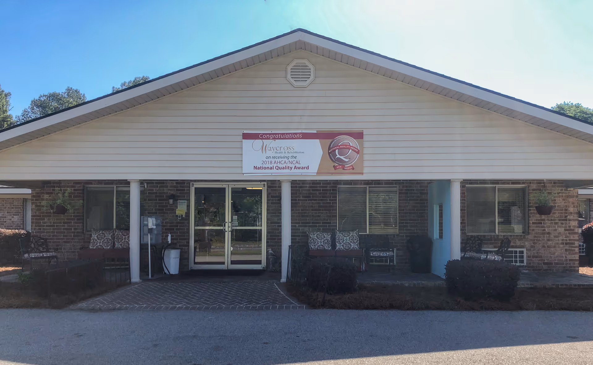 Front entrance of Waycross Health & Rehabilitation, a single-story brick building with a covered porch, glass double doors, benches, planters, and a sign above the entrance.