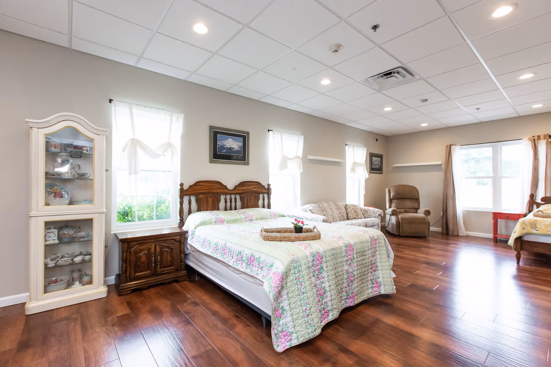 A spacious bedroom with wooden flooring featuring a double bed with a floral quilt, a wooden nightstand, a glass-front cabinet with decorative dishes, a floral-patterned sofa, a beige recliner chair, and large windows with sheer white curtains allowing natural light to fill the room.