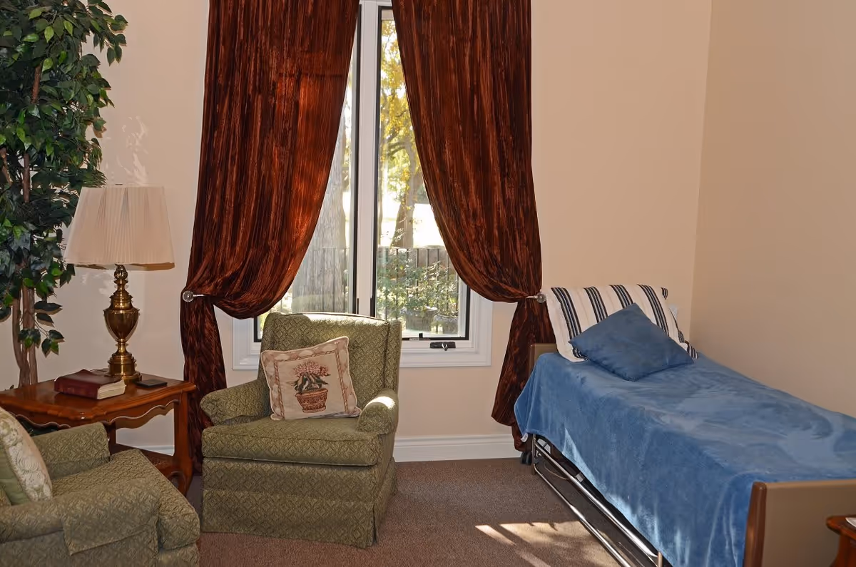 A cozy assisted-living bedroom with a single bed, upholstered armchairs, a side table and lamp, and a window with brown curtains.