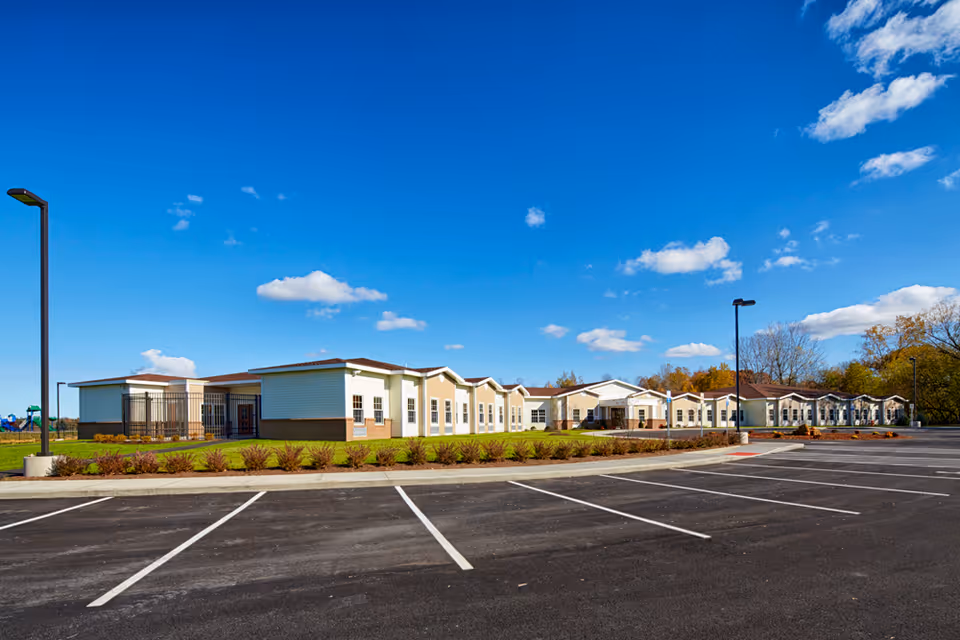 Wide exterior view of St. Francis Commons Assisted Living Residence on a clear sunny day with a bright blue sky and scattered clouds. The building is single-story with light-colored siding and a brown roof. In front of the building is a large empty parking lot with marked parking spaces and several tall street lamps. There are landscaped areas with small bushes along the building and some trees in the background.