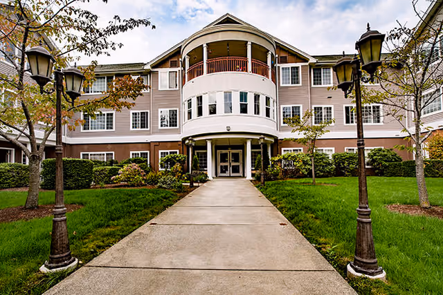 Front entrance of a three-story senior living building with a circular central portico, walkway, lamp posts, and manicured lawns.
